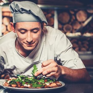 Food concept. Preparing traditional italian pizza. Young smiling chef in white uniform and gray hat decorate ready dish with green rucola herbs in interior of modern restaurant kitchen. Ready to eat.